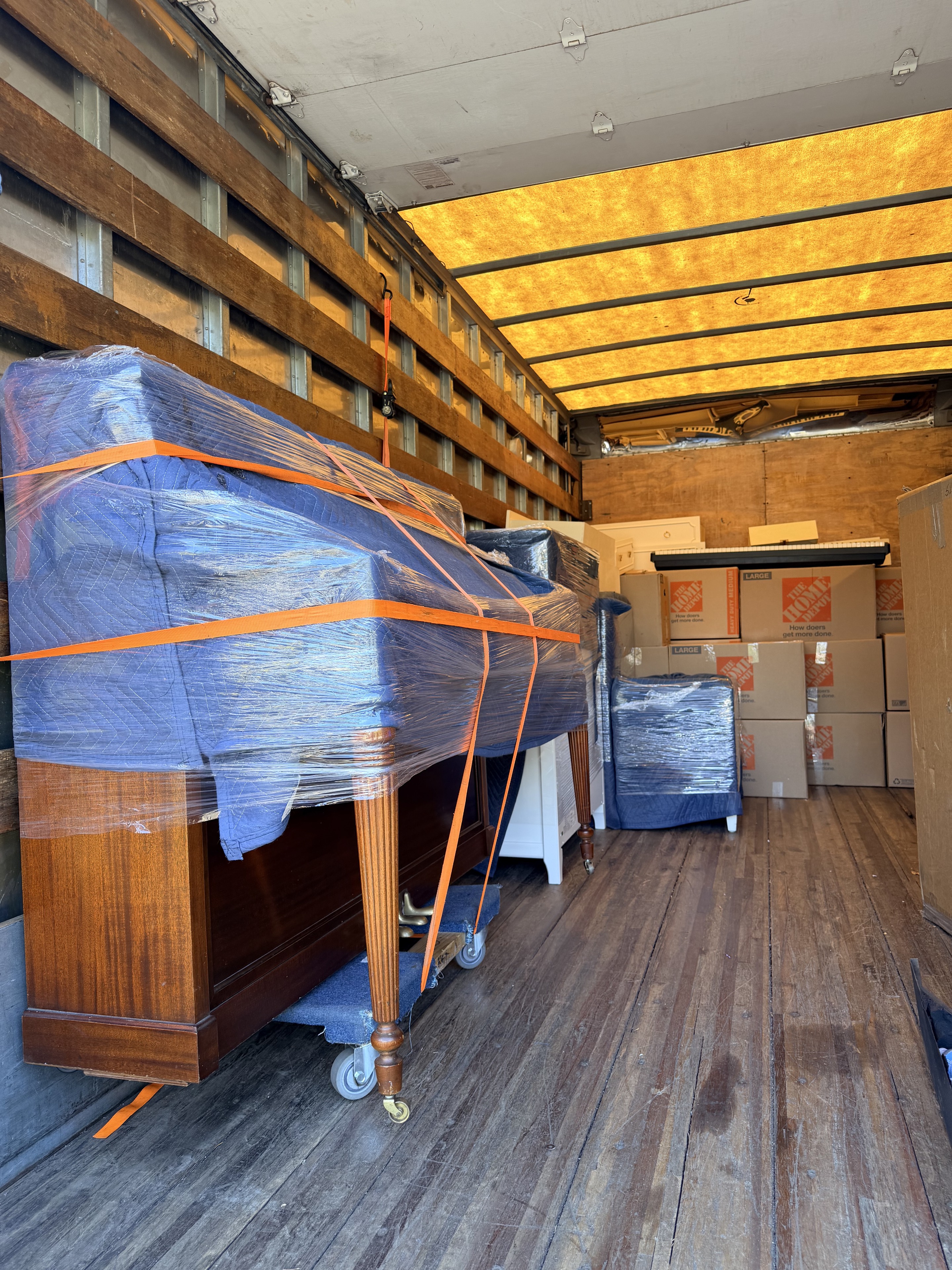 A piano protected with moving blankets and secured inside a moving truck with orange straps.