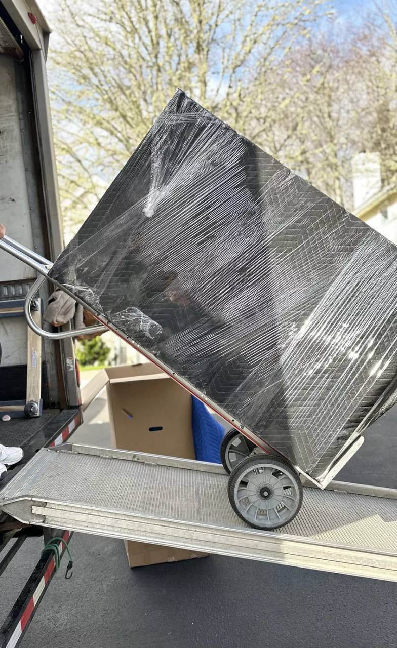 A large wrapped furniture piece is guided on a hand truck at the moving truck ramp.